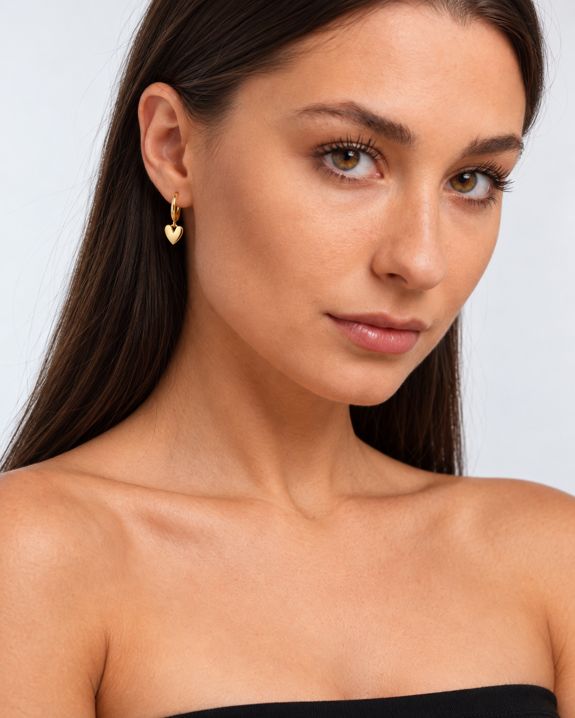 Small gold-coloured hoop earrings with a hanging heart worn by a brunette model, three-quarter view.